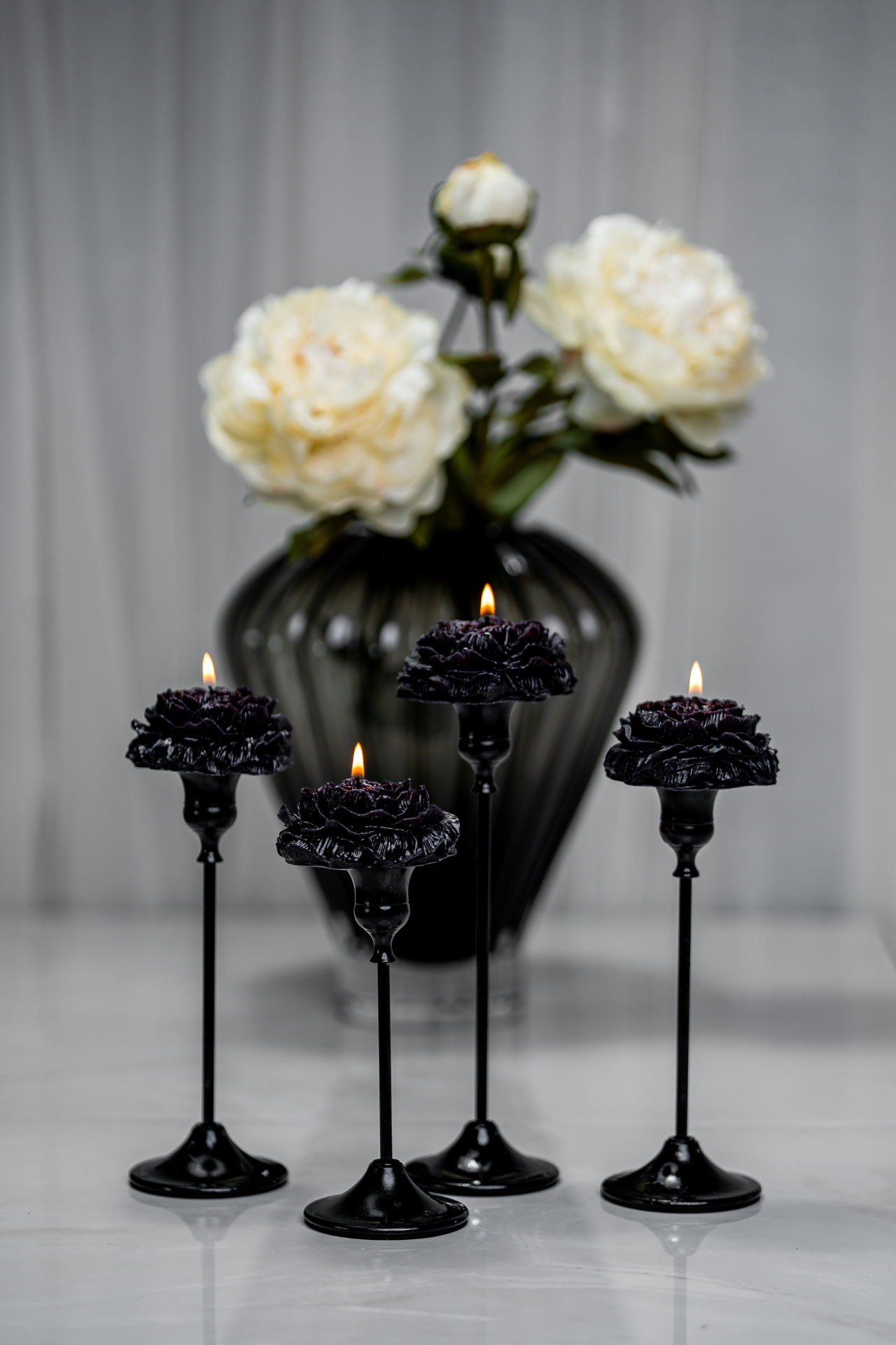 Four black PetiteBlooms in black candle holders, lit with warm flames, styled in front of white peonies